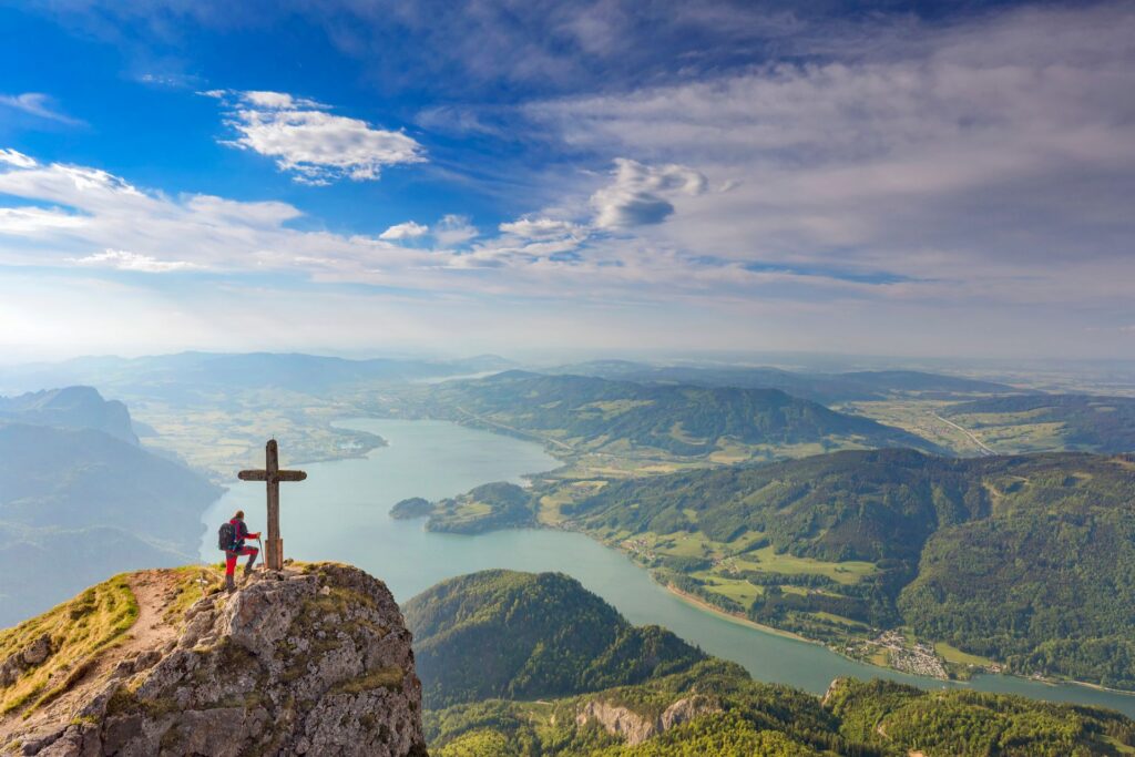 Eine Person steht neben einem Gipfelkreuz auf einem Felsen und blickt auf einen weitläufigen See sowie hügelige, grüne Landschaften unter einem teils bewölkten Himmel.