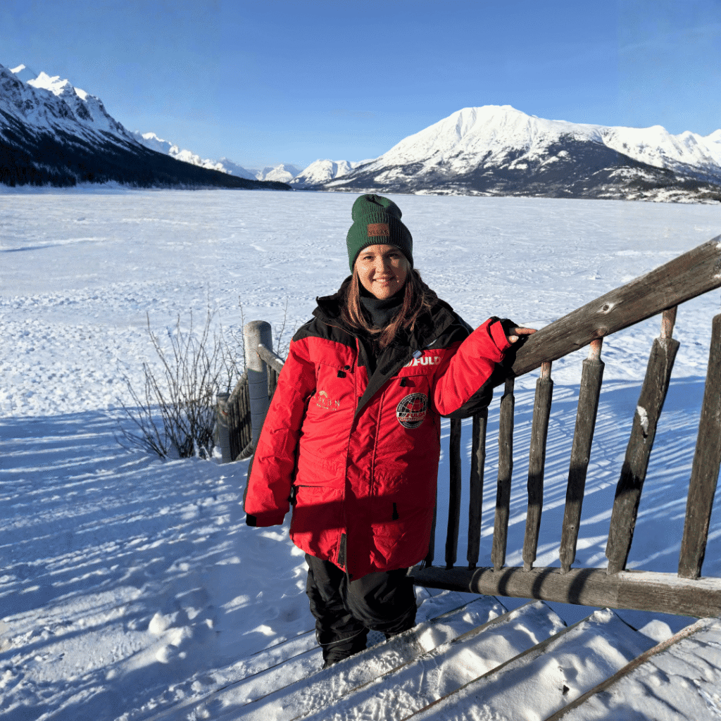 Person in roter Winterjacke vor zugefrorenem See. Zugefrorener See in kanadischer Winterlandschaft mit schneebedeckten Bergen und klarer Sonne.