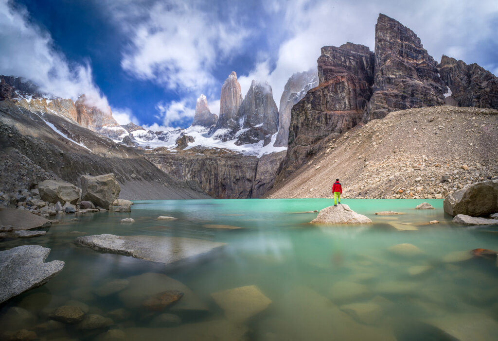 Ein Wanderer mit roter Jacke steht auf einem Stein im türkisfarbenden Bergsee und blickt auf drei markante Bergspitzen im Hintergrund.