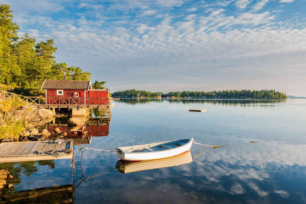 Urlaub in Schweden - Eine kleine rote Holzhütte liegt am Rande eines ruhigen Sees, perfekt für einen friedlichen Urlaub in Schweden. Ein weißes Ruderboot liegt am Steg, während Bäume und ein teilweise bewölkter Himmel diese malerische schwedische Landschaft vervollständigen.