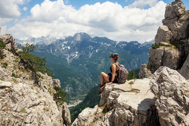 Wanderin sitzt auf Stein und blickt in die Ferne. Die Fernlandschaft sind teilweise schneebedeckte Berge.
