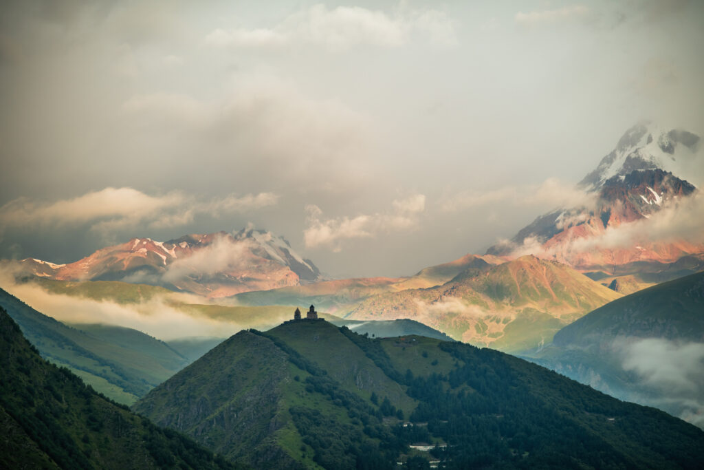 Berglandschaft mit Kirche im Vordergrund auf Berg. Im Hintergrund leuchtende Berge umgeben von Wolken.