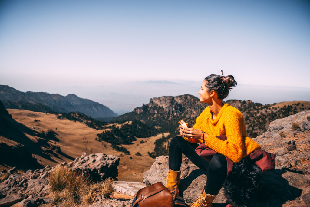 Person sitzt auf einem Felsen in einer Berglandschaft und blickt über ein weites Tal.