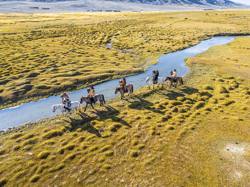 Gruppe von Reitenden überquert auf Pferden einen schmalen Fluss in einer weiten Graslandschaft.