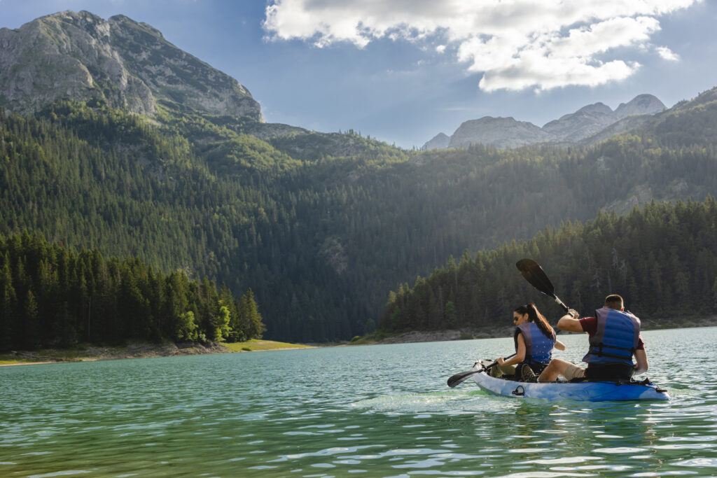 Zwei Personen paddeln gemeinsam in einem Schlauchboot auf einem türkisfarbenen Bergsee, umgeben von bewaldeten Hängen und felsigen Bergen unter einem teils bewölkten Himmel.