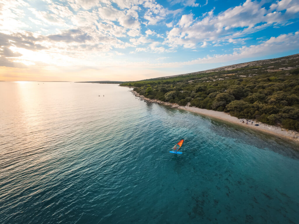 Luftaufnahme eines älteren Mannes beim Windsurfen auf ruhigem, klarem, blauem Wasser. Die Aufnahme, die direkt von oben gemacht wurde, hebt das leuchtende Segel, die Bewegung des Boards und die Weite des offenen Meeres hervor. Der Mann wirkt aktiv und konzentriert und verkörpert Abenteuerlust, Vitalität und Freiheit. Ideal für Themen wie gesundes Altern, Sport, Lifestyle, Reisen und Outdoor-Aktivitäten. Aufgenommen in der Nähe der Insel Pag in Kroatien in der Adria.