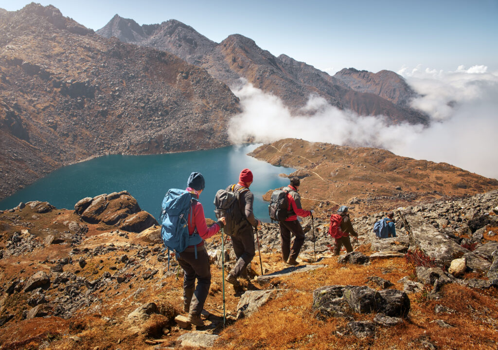 Gruppe von Wandernden mit Rucksäcken steigt auf einem felsigen Bergpfad oberhalb eines türkisfarbenen Bergsees in einer kargen Hochgebirgslandschaft ab.