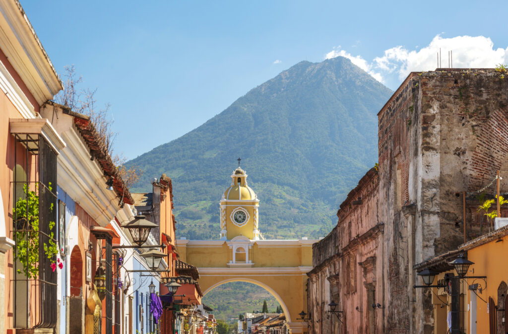 Kolonialstraße in Antigua Guatemala mit gelbem Bogen und Uhr sowie Vulkan im Hintergrund unter blauem Himmel.
