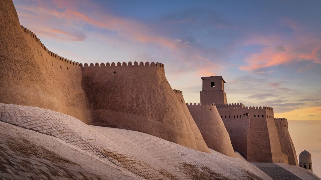 Massive Lehmziegel-Stadtmauern der Altstadt von Chiwa (Itchan Kala) mit Zinnen und Wachturm im warmen Licht eines farbenfrohen Sonnenuntergangs in Usbekistan.