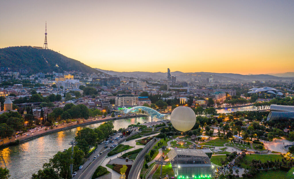 Panoramablick bei Sonnenuntergang auf eine Stadt mit Fluss, moderner Glasbrücke, Parkanlage und Sendeturm auf einem Hügel im Hintergrund.