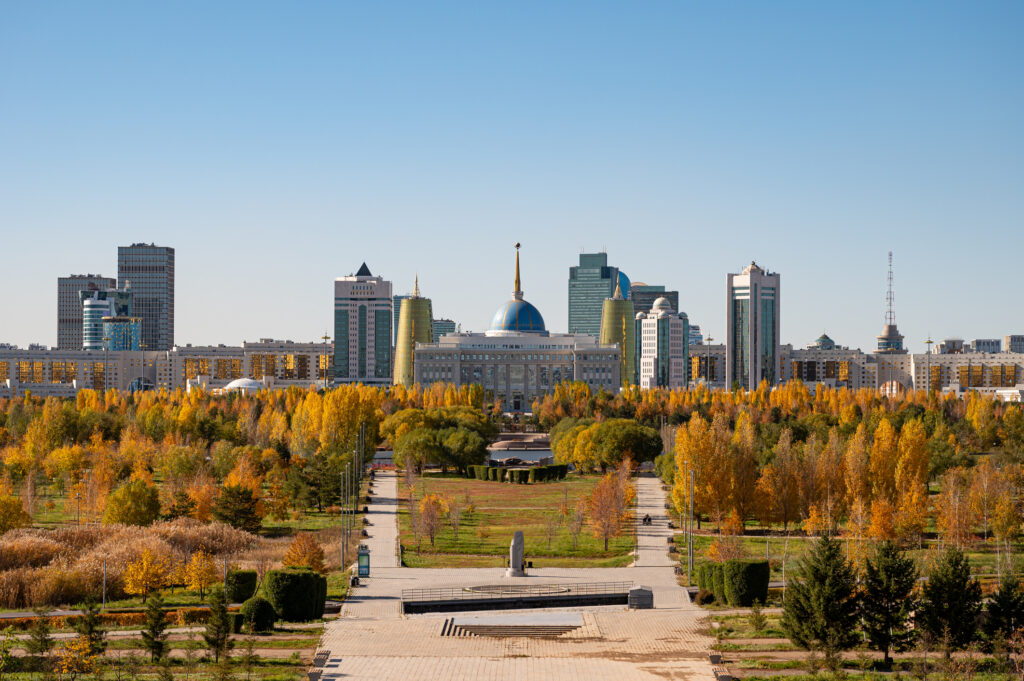 Stadtansicht mit moderner Skyline, zentralem Kuppelgebäude und großem Park mit herbstlich gefärbten Bäumen im Vordergrund.