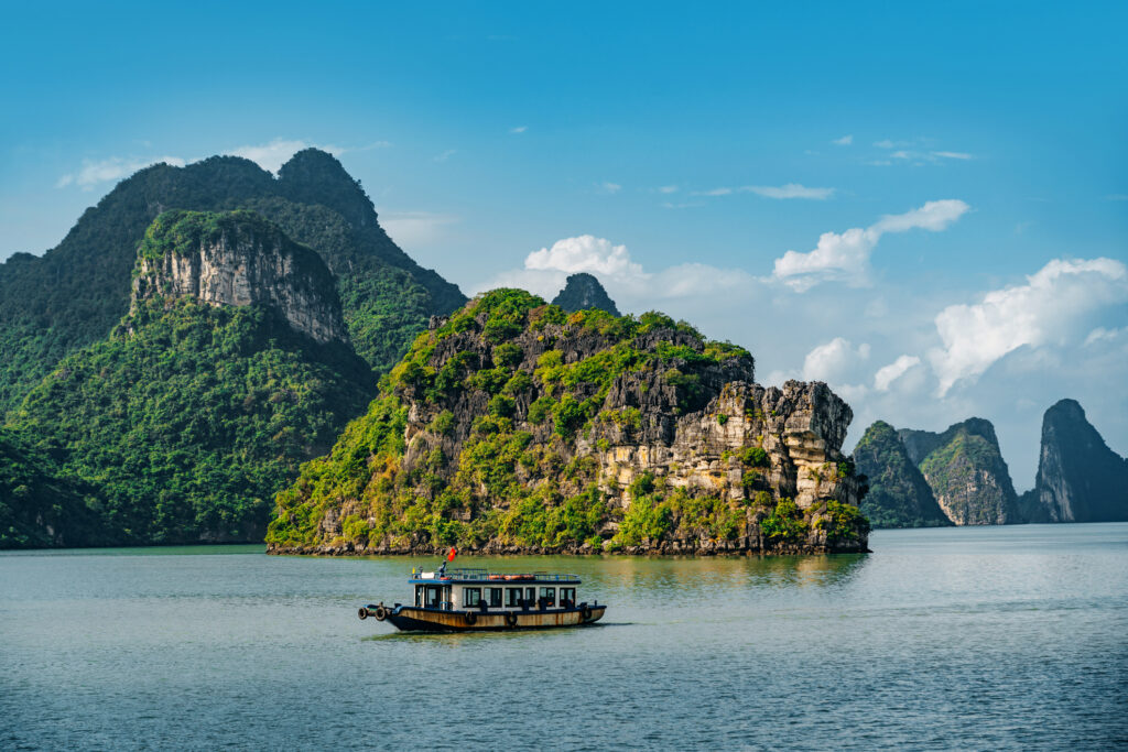 Traditionelles Boot auf ruhigem Wasser in der Halong-Bucht, umgeben von üppig bewachsenen Kalksteinfelsen unter einem klaren blauen Himmel in Vietnam.
