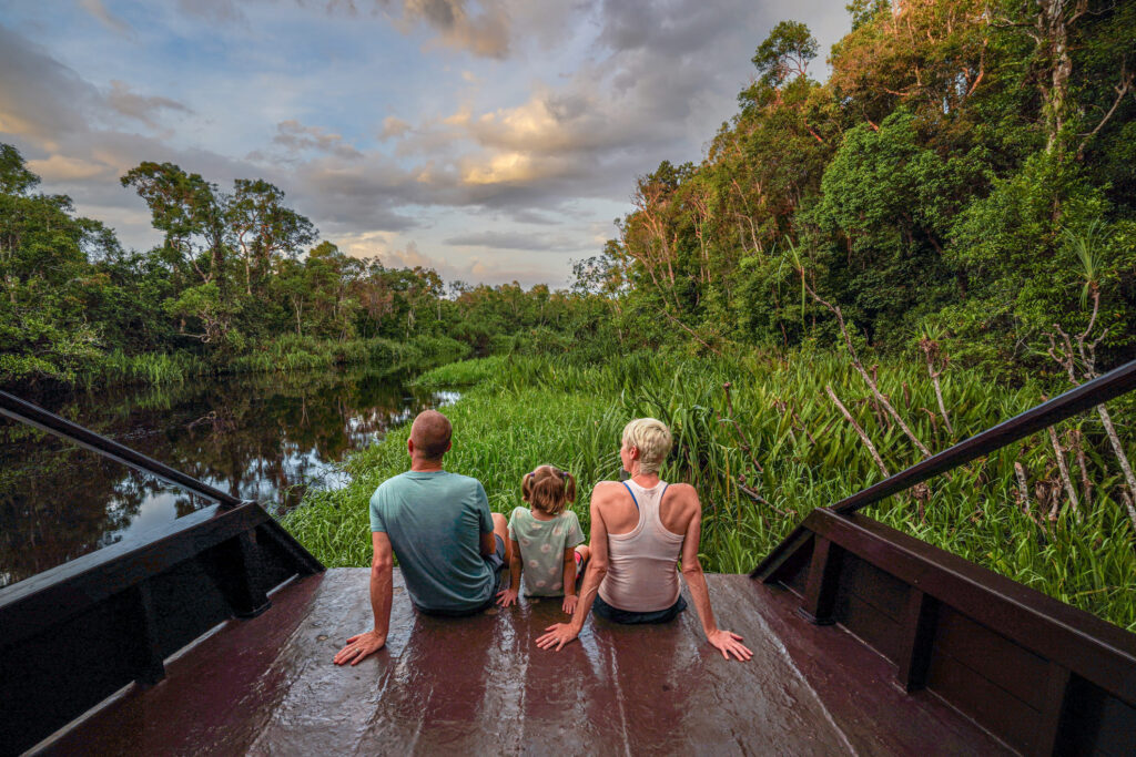 Eine Familie sitzt auf dem Bug eines Flussboots und blickt bei Sonnenuntergang auf den dschungelgesäumten Kinabatangan-Fluss in Borneo.