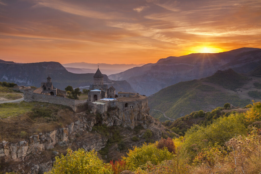 Tatev-Kloster in Armenien auf einem Felsen über einer tiefen Schlucht, umgeben von Bergen und herbstlicher Vegetation, bei einem leuchtenden Sonnenuntergang.