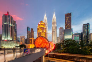 kyline von Kuala Lumpur zur blauen Stunde mit den beleuchteten Petronas Towers, einer beleuchteten runden Fußgängerbrücke im Vordergrund und der malaysischen Flagge am Geländer.