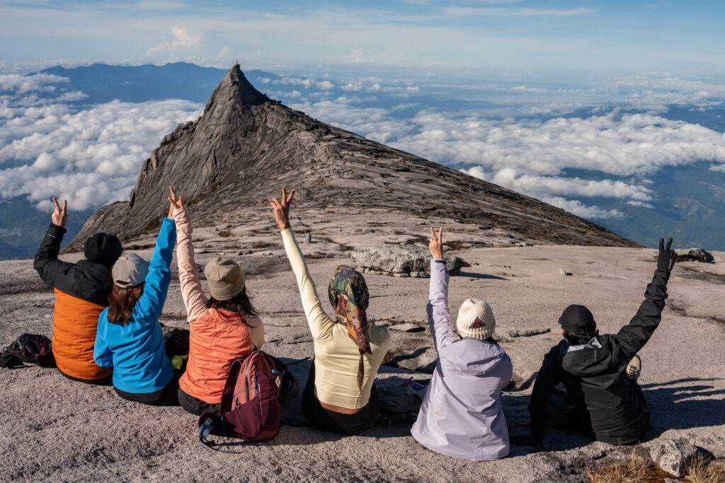 Eine Gruppe von Wanderern jubelt mit erhobenen Armen über einem Wolkenmeer nahe dem markanten Gipfel des Mount Kinabalu in Borneo.