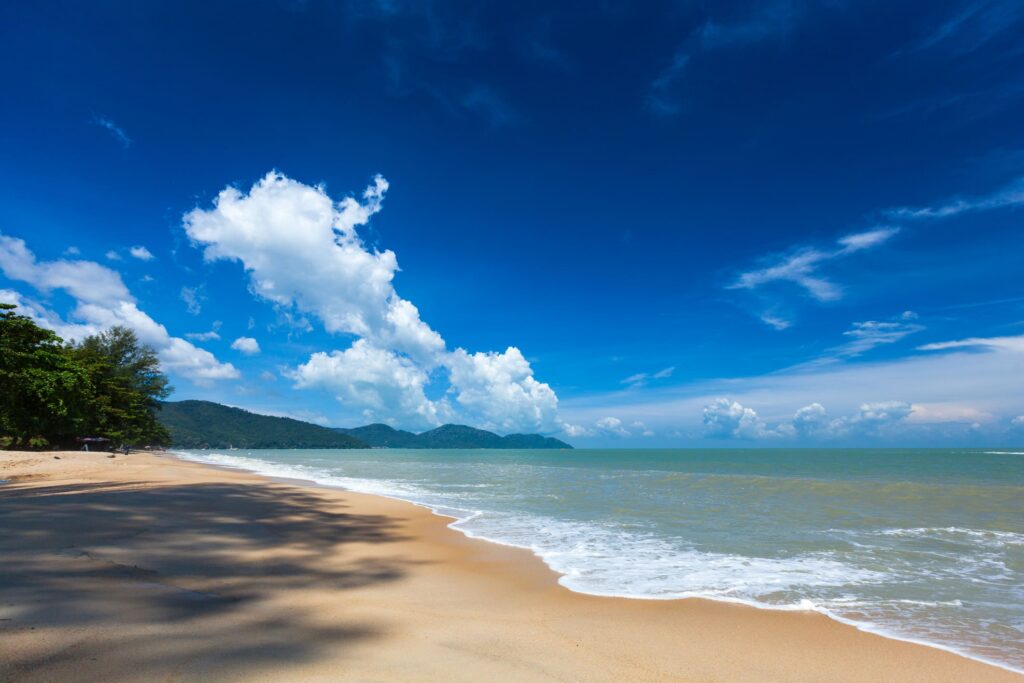 Ein langer, heller Sandstrand verläuft von links vorne bis in die Ferne entlang einer tropischen Küste. Rechts trifft sanftes, hellblaues Meerwasser auf den Strand und hinterlässt einen schmalen Saum aus weißem Schaum. Auf der linken Seite spenden große Bäume Schatten auf den Sand. Im Hintergrund sind mehrere bewaldete Hügel oder Inseln am Horizont zu sehen. Über der Landschaft erstreckt sich ein intensiv dunkelblauer Himmel mit auffälligen, hohen weißen Wolkenformationen. Das Bild vermittelt die Weite und Ruhe einer tropischen Küstenlandschaft.