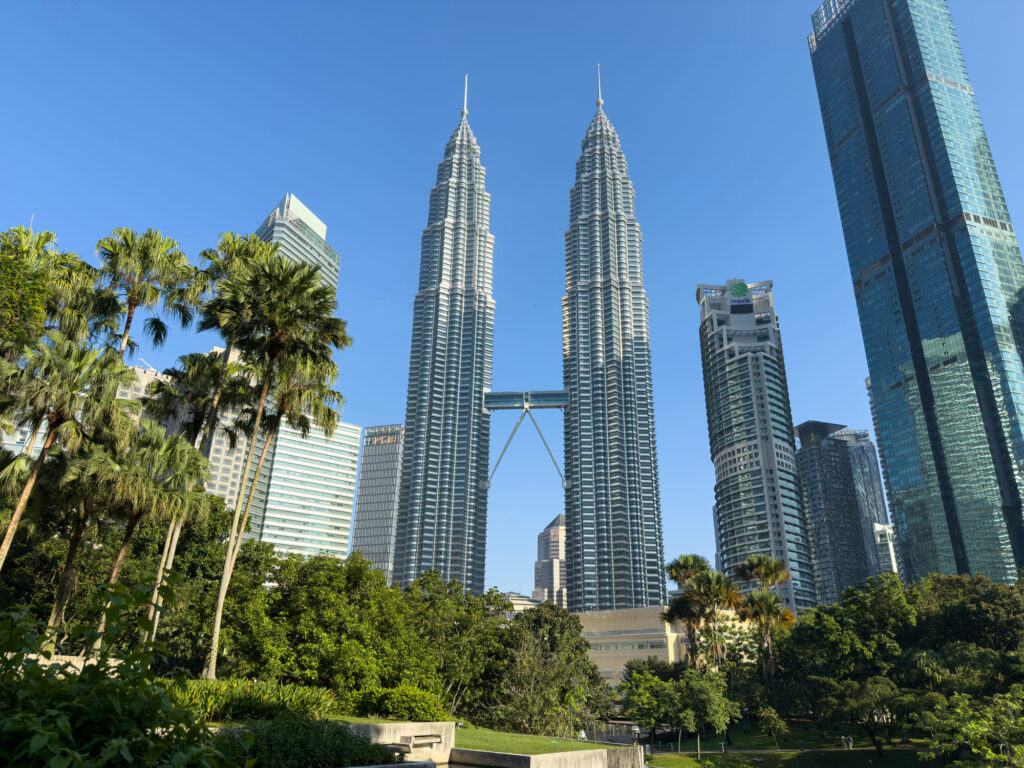 Die Petronas Towers in Kuala Lumpur ragen bei strahlend blauem Himmel zwischen Palmen und umliegenden Wolkenkratzern empor.