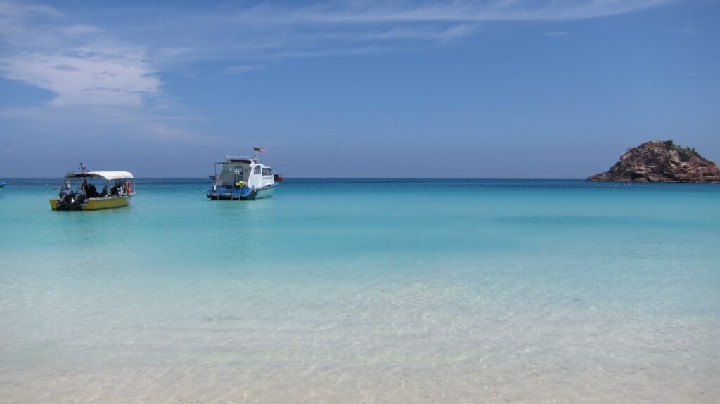 Zwei kleine Boote liegen auf ruhigem, kristallklarem türkisfarbenem Wasser nahe der Küste. Das flache Wasser im Vordergrund ist so klar, dass der helle Sandboden sichtbar bleibt. Am rechten Bildrand erhebt sich eine kleine felsige Insel aus dem Meer. Der Horizont trennt das dunklere Blau des offenen Meeres vom helleren Wasser der Lagune. Über der Szene spannt sich ein weiter Himmel mit dünnen Wolken. Das Bild vermittelt die Ruhe und Klarheit einer tropischen Inselbucht.