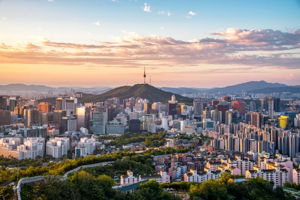 Panoramablick auf die Skyline von Seoul, Südkorea, bei Sonnenuntergang mit dem Namsan Seoul Tower auf einem bewaldeten Hügel inmitten dichter Hochhausbebauung und Bergen im Hintergrund.