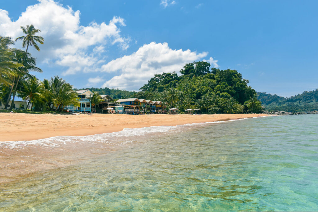Ein breiter Sandstrand erstreckt sich entlang einer tropischen Bucht. Im Vordergrund ist das klare, flache Meer zu sehen, dessen leicht türkisfarbenes Wasser sanft an den hellen Sand strandet. Entlang der Küste stehen mehrere Gebäude und kleine Unterkünfte direkt hinter dem Strand, eingerahmt von hohen Palmen und dichter tropischer Vegetation. Dahinter erhebt sich ein bewaldeter Hügel mit üppigem Grün. Der Himmel ist strahlend blau und mit großen, weißen Wolken bedeckt. Die Küstenlinie verläuft nach rechts in die Ferne, wo weitere bewaldete Hänge sichtbar sind. Das Bild vermittelt den Eindruck eines tropischen Inselortes, in dem Natur und touristische Infrastruktur harmonisch nebeneinander bestehen.