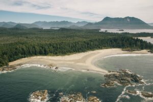 Luftaufnahme einer geschwungenen Sandbucht mit hellem Strand, dichtem Küstenwald und felsigen Uferabschnitten. Im Hintergrund erstrecken sich Inseln und Berge entlang der Küste. Die Szene zeigt die Weite und Ursprünglichkeit einer naturbelassenen Küstenlandschaft.