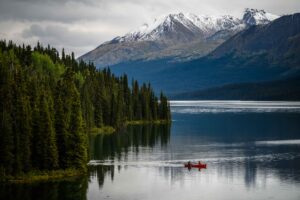 Ein rotes Kanu mit zwei Personen gleitet über einen ruhigen Bergsee. Dichte Nadelwälder säumen das Ufer, während sich im Hintergrund schneebedeckte Berge über dem Wasser erheben. Die spiegelnde Oberfläche des Sees unterstreicht die Ruhe und Weite der Landschaft.