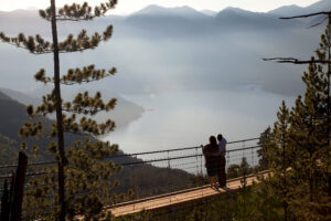 Zwei Personen stehen auf einer Aussichtsplattform hoch über einem See und blicken auf die umliegenden Berge. Hohe Kiefern rahmen die Szene ein, während das warme Sonnenlicht die Landschaft in einen leichten Dunst hüllt.