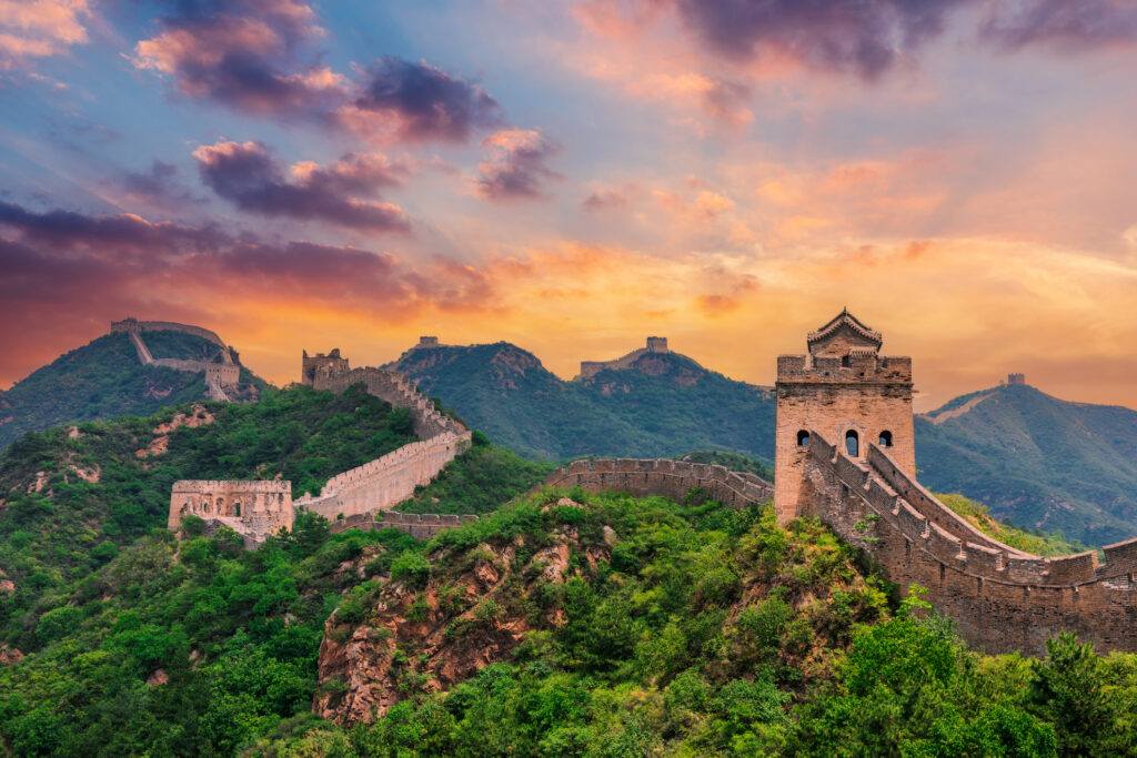 Chinesische Mauer mit Wachtürmen auf bewaldeten Hügeln in China bei spektakulärem Sonnenuntergang mit farbenfrohem Himmel.