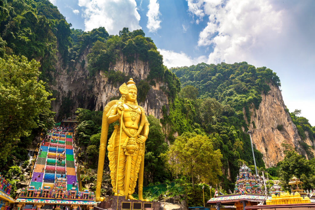 Goldene Murugan-Statue vor den bunten Treppen der Batu Caves in Malaysia, umgeben von Kalksteinfelsen, Tempelbauten und tropischer Vegetation.