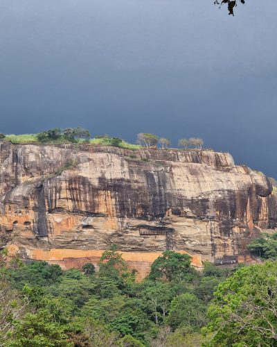 Sigiriya-Felsen in Sri Lanka mit dichter Vegetation und dunklen Regenwolken im Hintergrund.