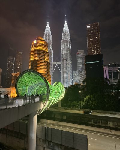 Beleuchtete Petronas Towers in Kuala Lumpur bei Nacht mit futuristischer grüner Fußgängerbrücke und moderner Skyline.