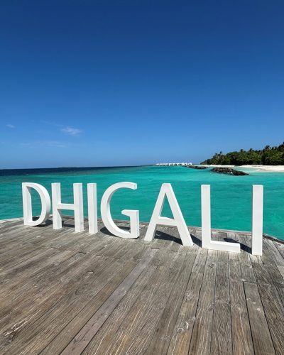 Weißer DHIGALI-Schriftzug auf Holzplattform vor türkisblauem Meer und tropischer Inselkulisse auf den Malediven.