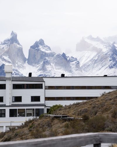 Weiße Lodge am Berghang mit schneebedeckten Gipfeln des Torres-del-Paine-Massivs im Hintergrund.
