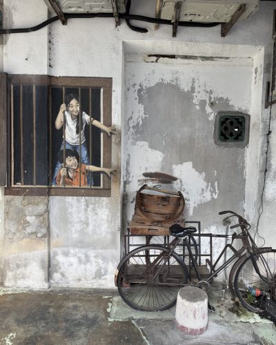 Street-Art-Motiv mit zwei Kindern an einer Hauswand in George Town, Penang, daneben ein altes Fahrrad und ein hölzerner Wagen.