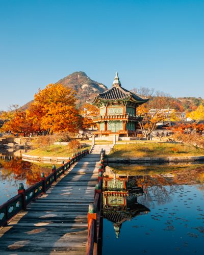 Hyangwonjeong-Pavillon im Gyeongbokgung-Palast in Seoul, Südkorea, mit hölzerner Brücke über einen Teich, umgeben von farbenfrohen Ahornbäumen im Herbst und spiegelnden Reflexionen im Wasser.