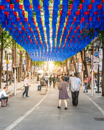 Belebte Fußgängerzone in Insa-dong, Seoul, mit zahlreichen Passanten, Geschäften und über der Straße aufgehängten blauen und roten Lampions zwischen schattenspendenden Bäumen.