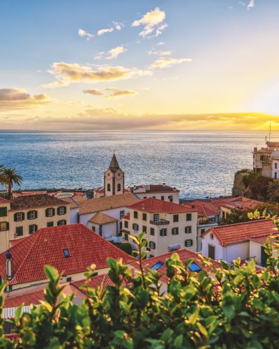 Blick auf Ponta do Sol auf Madeira bei Sonnenuntergang mit roten Dächern, Kirchturm, Atlantik und goldenen Sonnenstrahlen über der Küste.