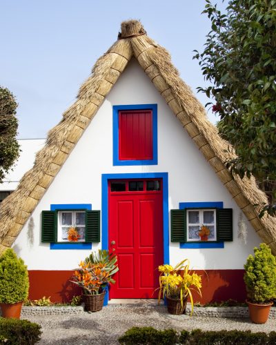 Traditionelles Santana-Haus auf Madeira mit strohgedecktem Dreiecksdach, weißen Wänden, roter Eingangstür, blau umrahmten Fenstern und dekorativen Pflanzen vor dem Gebäude.