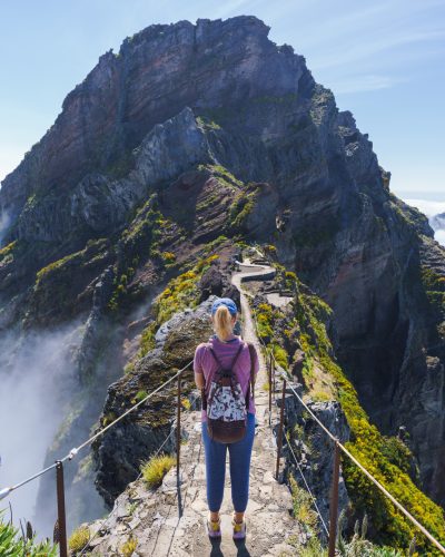 Wandernde Person auf dem PR1-Wanderweg zwischen Pico do Arieiro und Pico Ruivo auf Madeira, umgeben von Berggipfeln, Wolken und steilen Felsformationen.