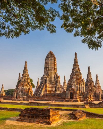 Alte Tempelruinen mit hohen Pagoden in Ayutthaya, Thailand, umgeben von Grünflächen und Bäumen im warmen Sonnenlicht.