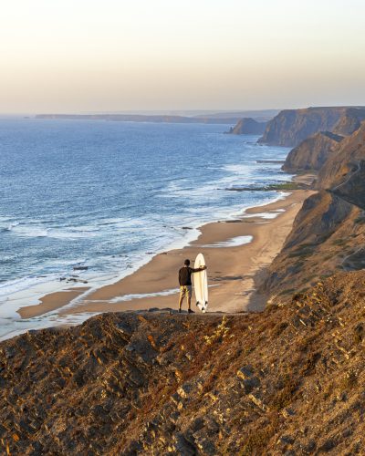 Rear view of a surfer with surfboard  standing on the edge of a cliff overlooking the ocean at sunset, Vila do Bispo, Sagres, Algarve, Portugal, Europe