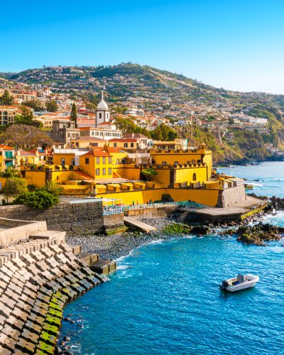 Panoramablick auf Funchal auf Madeira mit dem gelben Forte de São Tiago an der Küste, dicht bebauten Berghängen, Atlantik und einem kleinen Boot im türkisblauen Wasser.