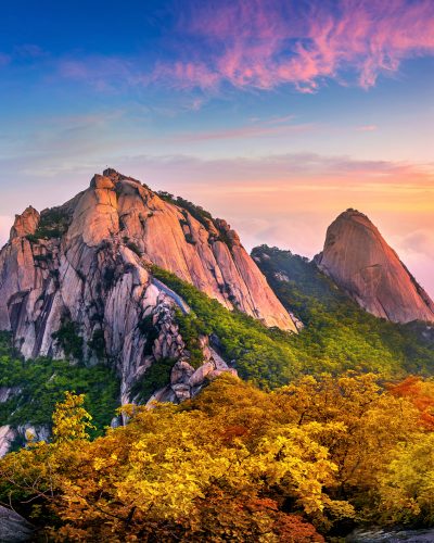 Granitgipfel des Bukhansan-Gebirges in Südkorea bei Sonnenaufgang mit bunten Herbstwäldern, Nebel zwischen den Bergen und einem farbenreichen Himmel in Rosa-, Blau- und Goldtönen.