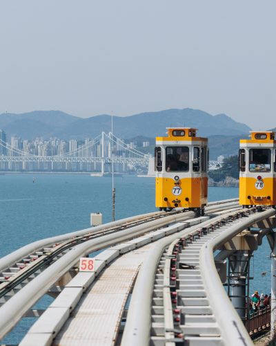 Zwei gelbe Sky-Capsule-Wagen des Haeundae Blue Line Parks fahren auf einer erhöhten Küstenbahn über dem Meer in Busan, mit Stadtpanorama, Brücke und Bergen im Hintergrund.