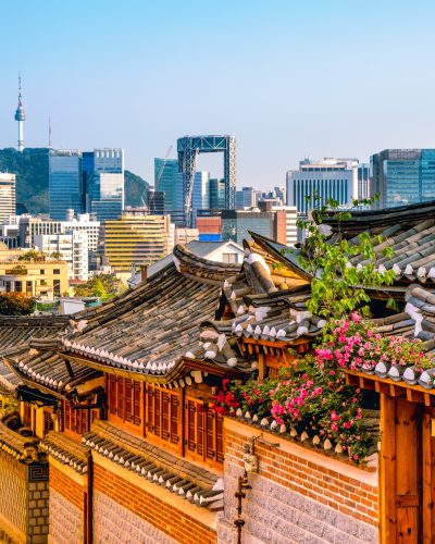 Traditionelle Hanok-Häuser im Bukchon Hanok Village in Seoul mit geschwungenen Ziegeldächern, blühenden Blumen und moderner Hochhauskulisse sowie dem Namsan Seoul Tower im Hintergrund.