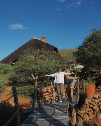 Person auf einem Holzsteg vor einer Safari-Lodge mit Reetdach im Tswalu Kalahari Reserve.