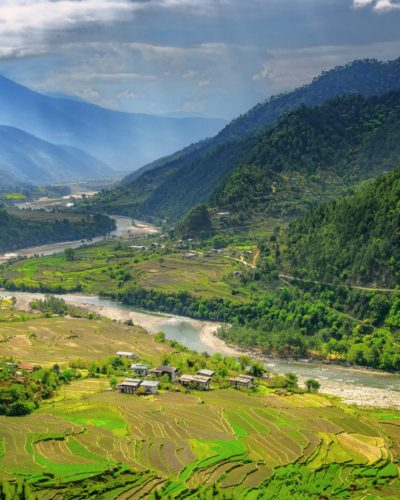 Klarer Bach in einer grünen Ebene des Gangtey-Tals in Bhutan, umgeben von Wiesen, bewaldeten Bergen, einzelnen Gebäuden und einem wolkenreichen Himmel.