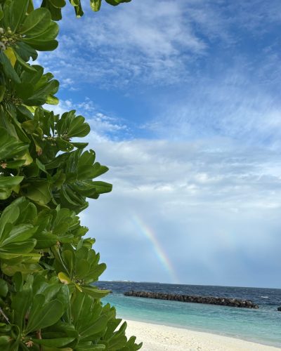 Regenbogen über einem Strand auf den Malediven mit tropischen Pflanzen, weißem Sand und türkisblauem Meer.