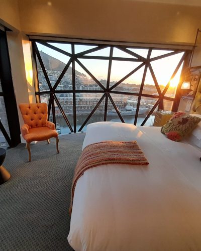 Modernes Hotelzimmer mit großem Bett und Panoramablick auf Hafen, Stadt und Sonnenuntergang.